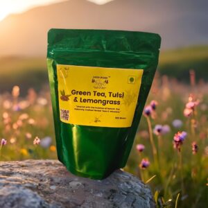 Front view of Jai Jungle Green Tea Tulsi Lemongrass loose tea 100g pack placed on a rock with wildflowers at sunset, highlighting natural herbal blend.